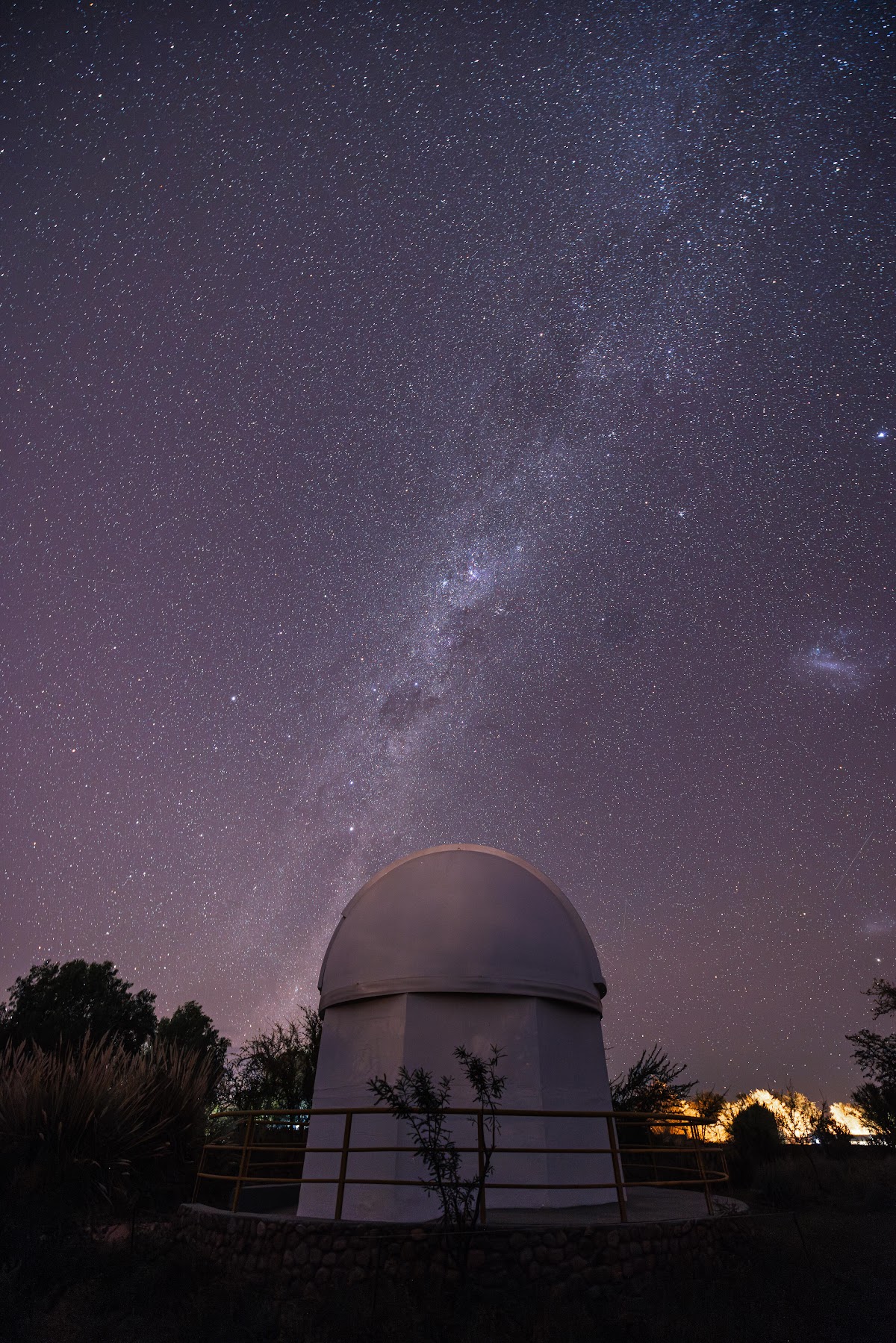 Observatory dome beneath the Milky Way at Explora Atacama, with desert vegetation and distant resort lights visible.