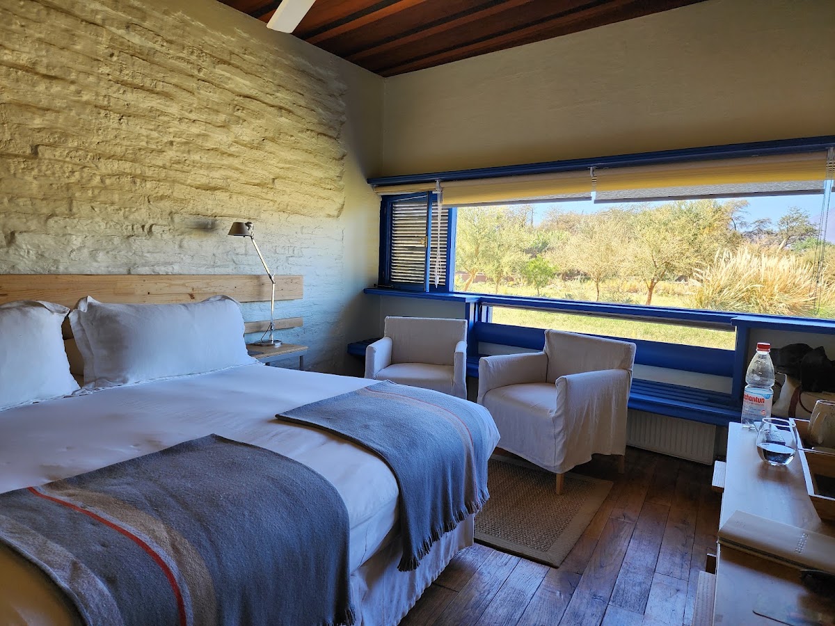 Hotel room with bed, seating area, and large windows overlooking desert landscape with sparse vegetation at Explora Atacama.