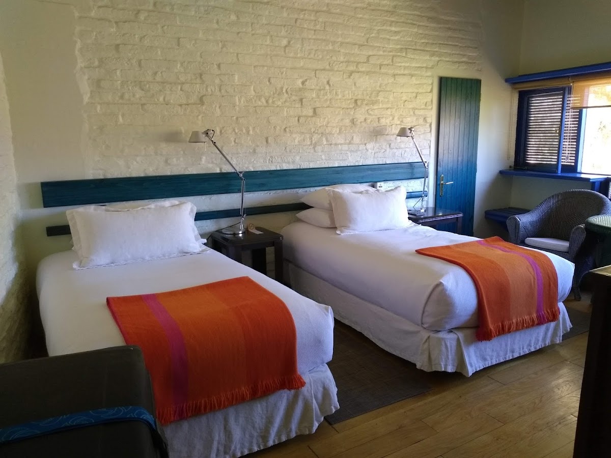 Resort bedroom with two twin beds topped with white linens and orange blankets, teal headboard, brick walls, and blue-trimmed windows and door.