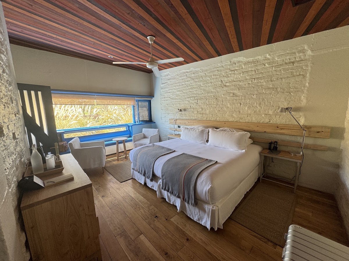 Spacious resort bedroom with wooden beam ceiling, queen bed with gray blanket, white brick accent wall, and blue-framed window overlooking desert landscape.