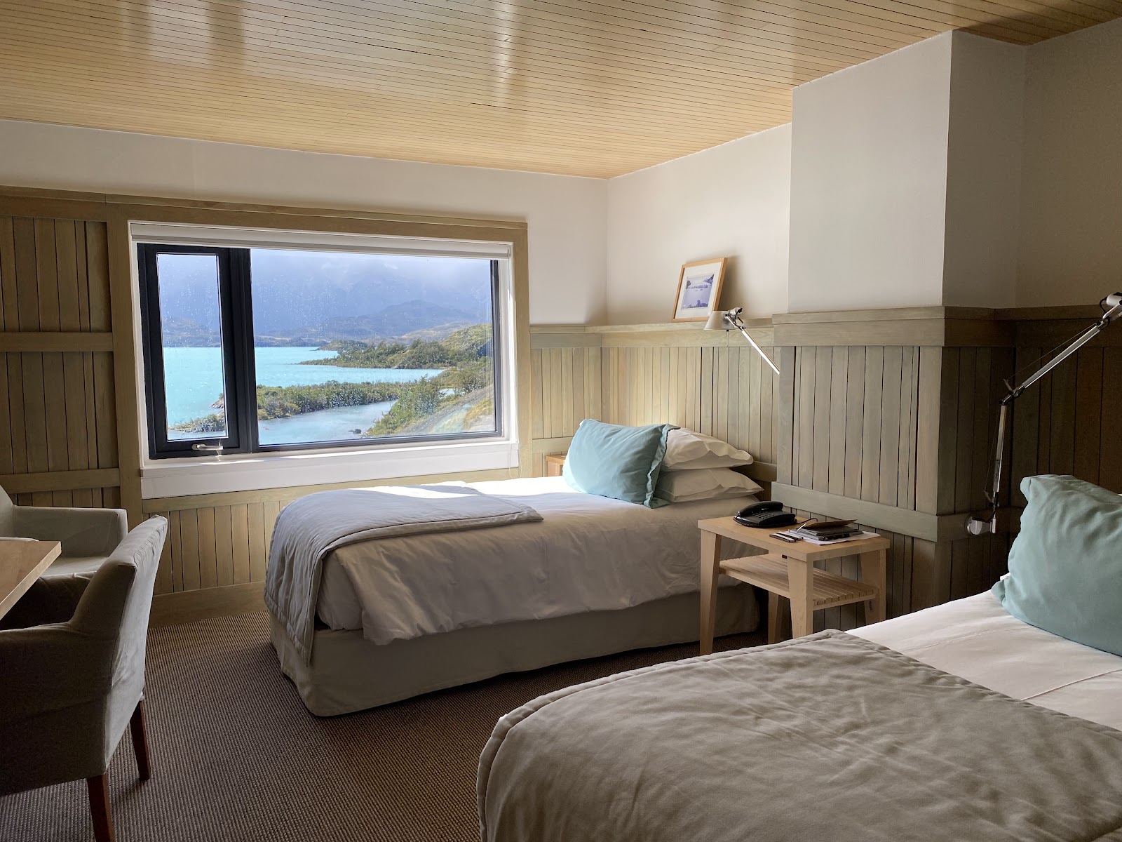 Resort room with two beds, wooden paneling, and large window overlooking turquoise lake and mountains at Explora Torres del Paine.