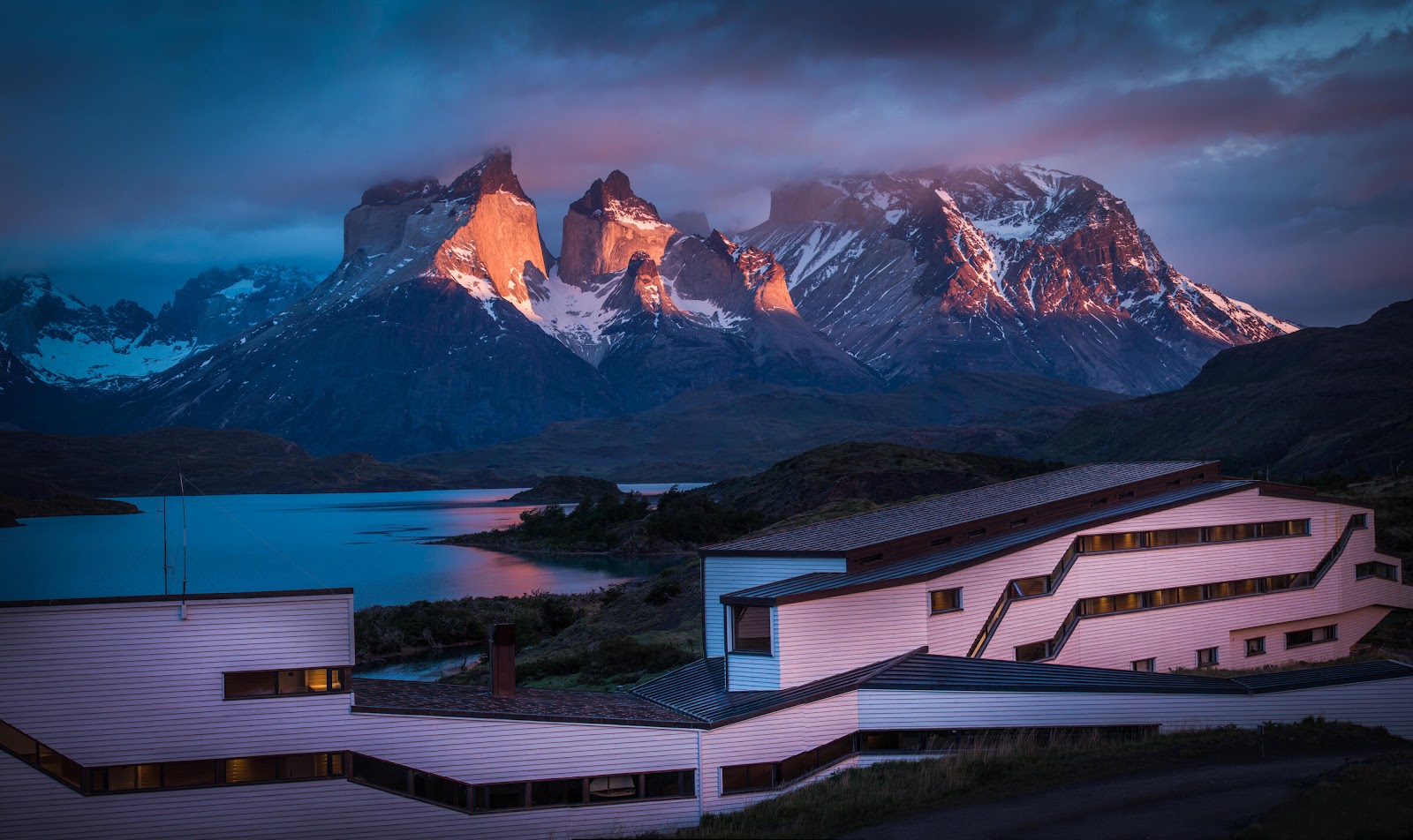 Modern white buildings with lit windows overlook a lake and snow-capped mountain peaks at dusk at Explora Torres del Paine.