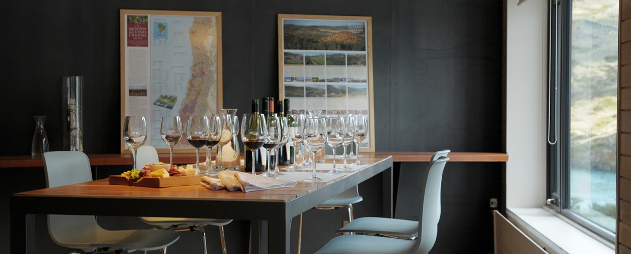 Wine tasting room at Explora Torres del Paine with lined glasses, cheese and bread on wooden counter, regional maps on dark walls, and mountain views through windows.