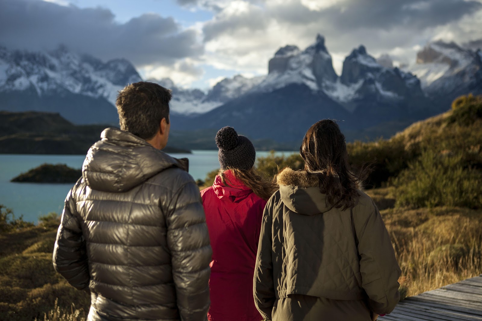 Three people in winter jackets view snow-capped mountain peaks and turquoise lake from a wooden overlook in Patagonia.