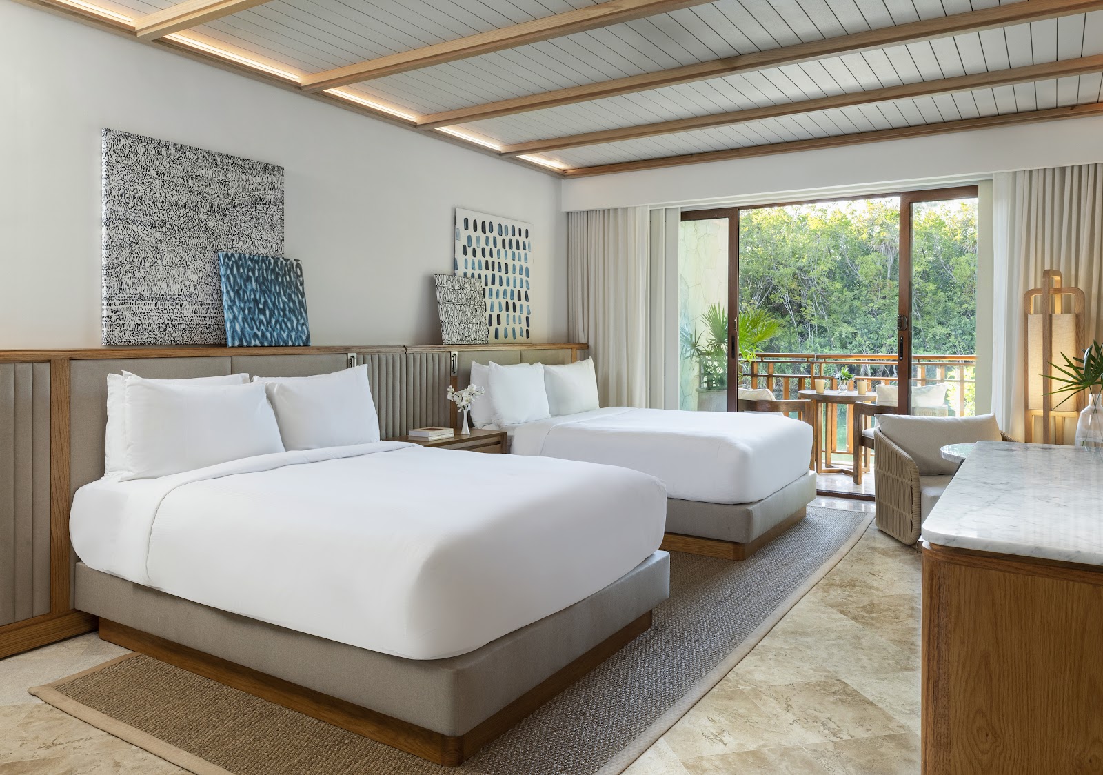 Modern resort suite with two queen beds, wood headboards, white linens, and glass doors opening to a forest-view balcony.