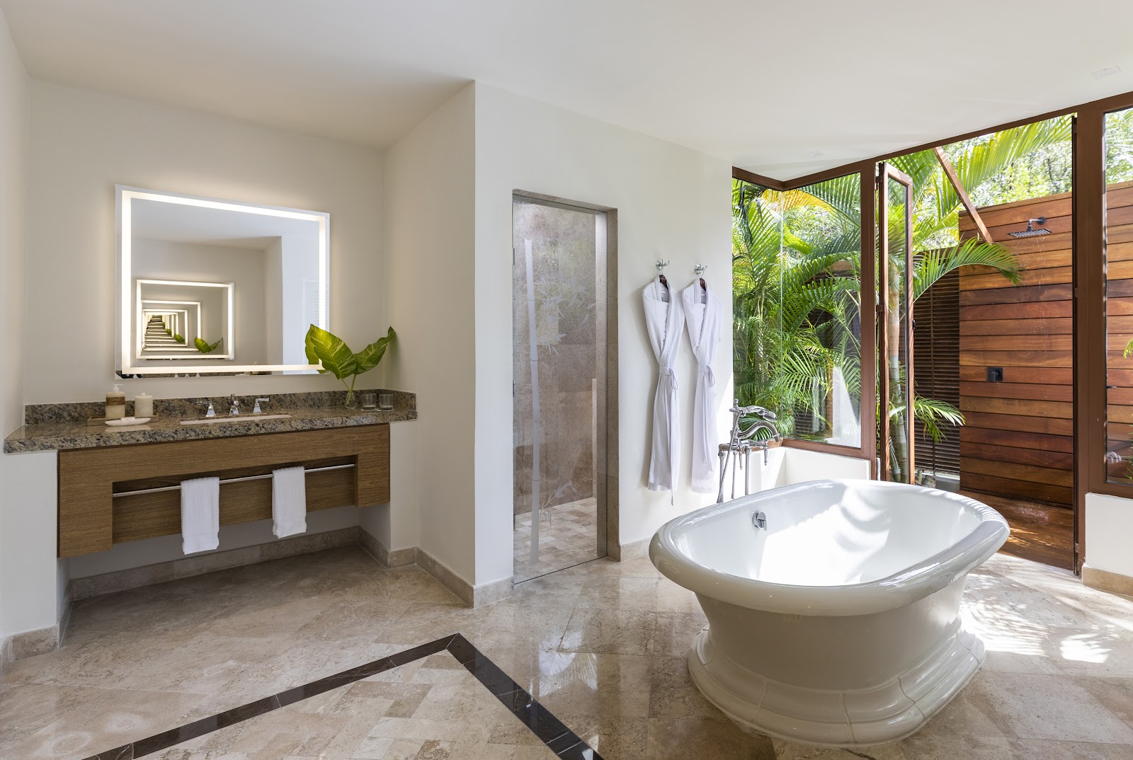 Spacious bathroom with freestanding oval tub, marble countertops, lighted mirror, and open doors to tropical garden views.