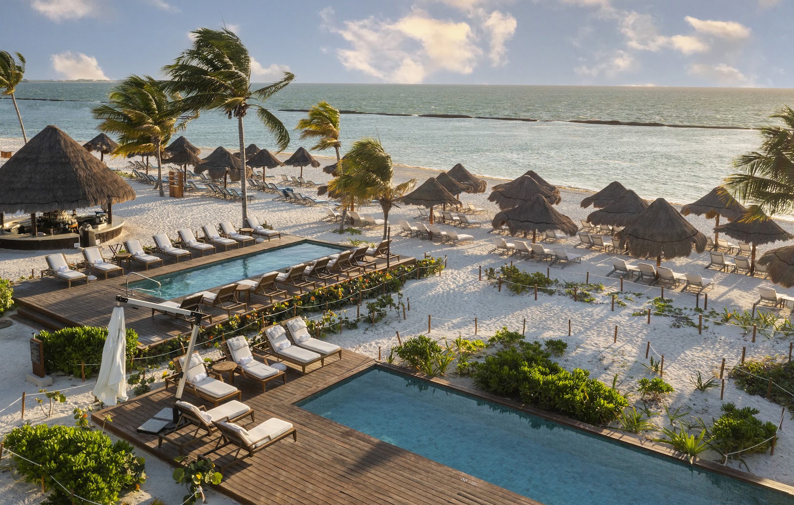 Resort pools and beach with thatched palapas, lounge chairs, palm trees, and turquoise water under partly cloudy sky.