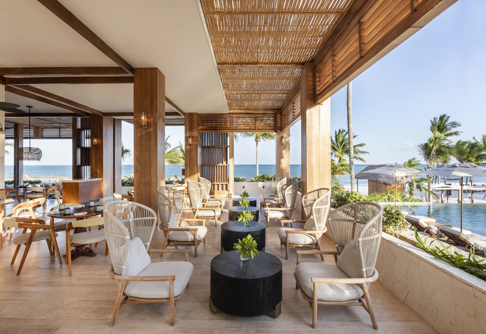 Covered terrace at Fairmont Mayakoba with woven ceiling, white wicker chairs, and ocean views beyond palm trees.