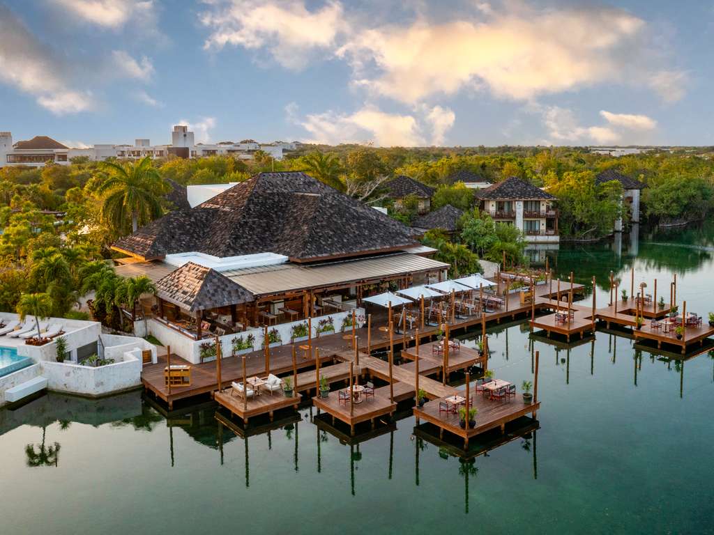 Fairmont Mayakoba's open-air lobby features cylindrical pendant lights, potted palms, round stone tables with wooden chairs, and a decorative bar with blue leopard sculptures in the background.