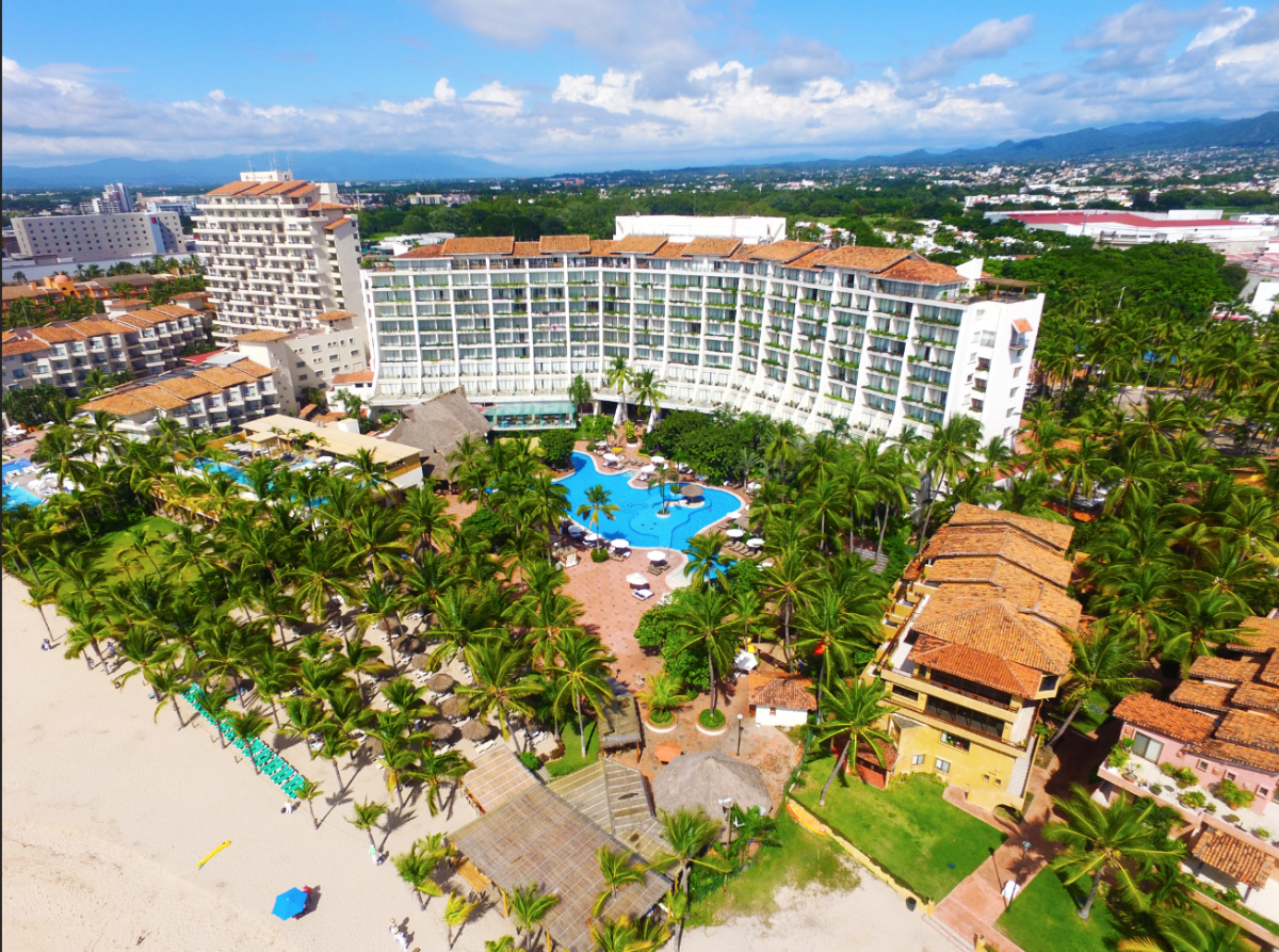 Aerial view of Fiesta Americana Puerto Vallarta showing beachfront resort with blue pool, palm trees, sandy beach, and mountain backdrop.