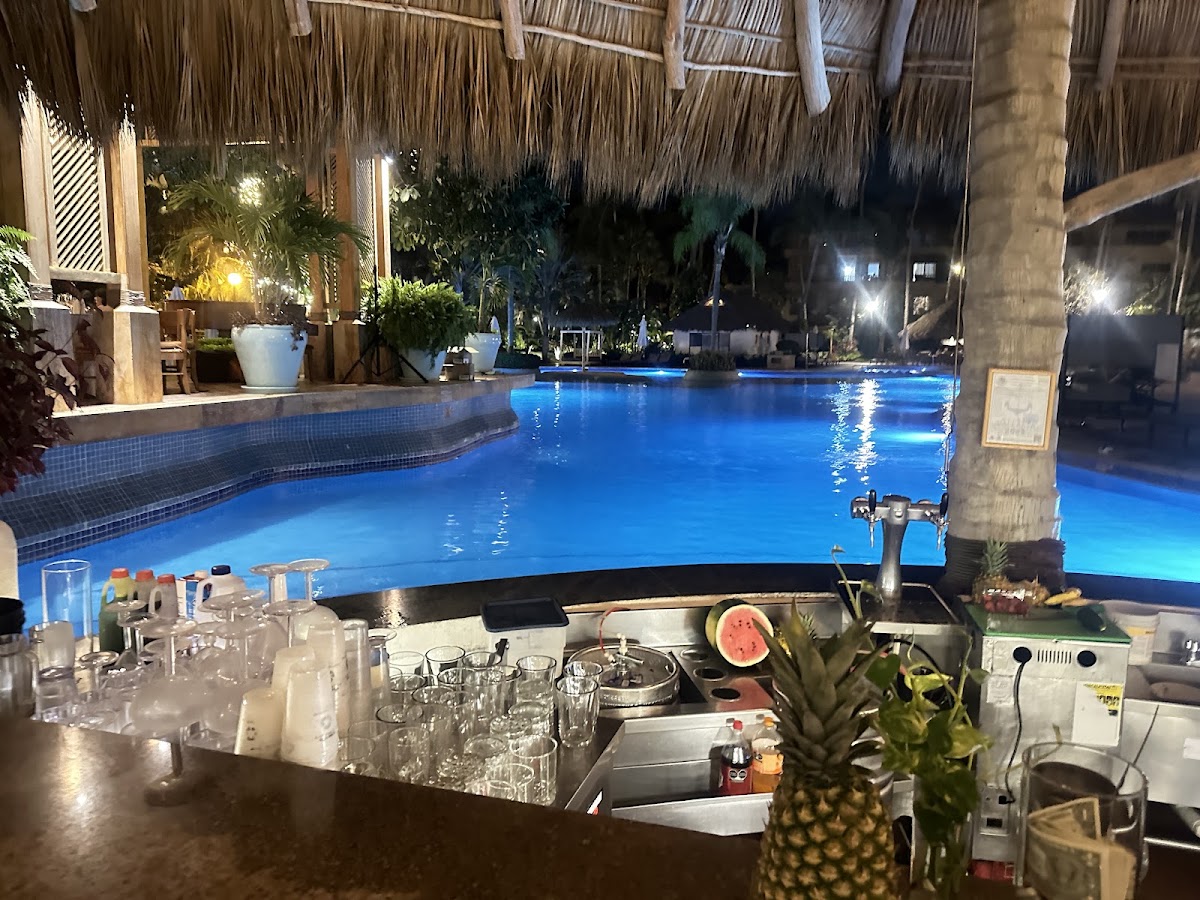Nighttime view of an illuminated blue pool with swim-up bar, thatched palapas, potted plants, and resort buildings under evening lighting at Fiesta Americana Puerto Vallarta.