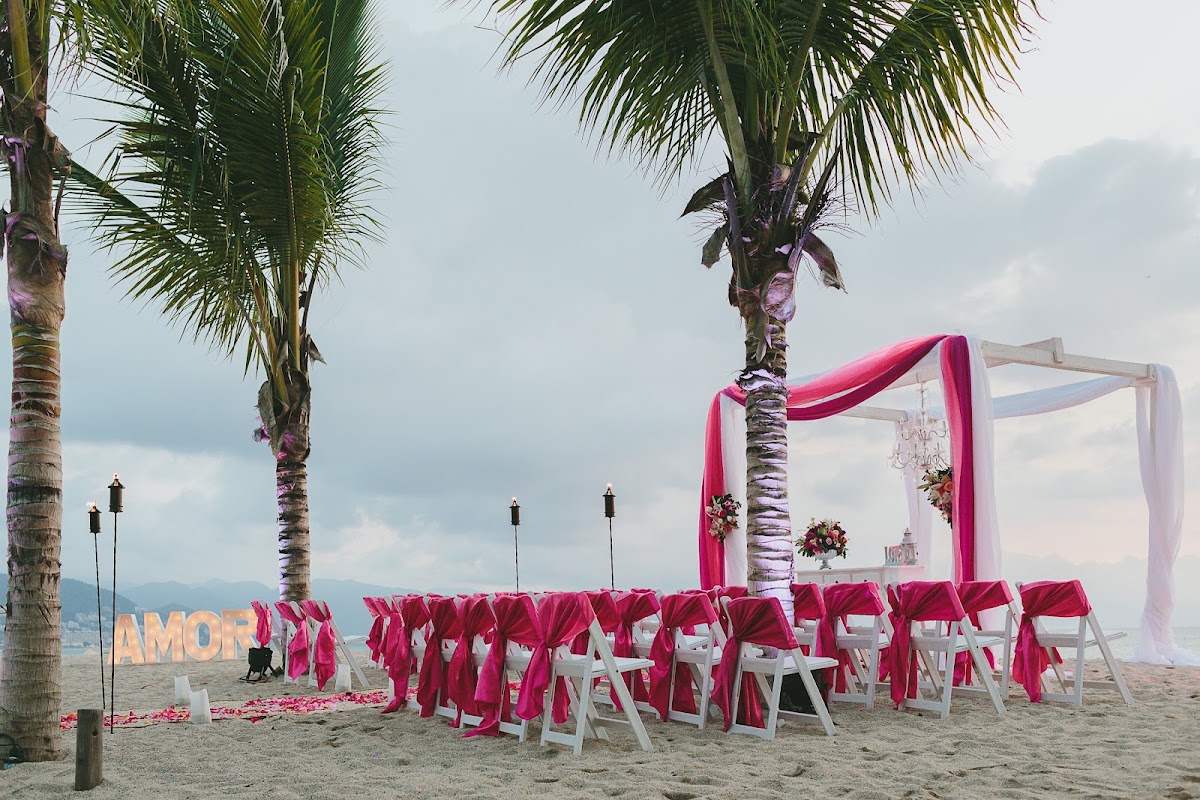 Beach wedding setup at Fiesta Americana Puerto Vallarta with white chairs draped in hot pink fabric, a pink and white arch, palm trees, and an AMOR sign on the sand.