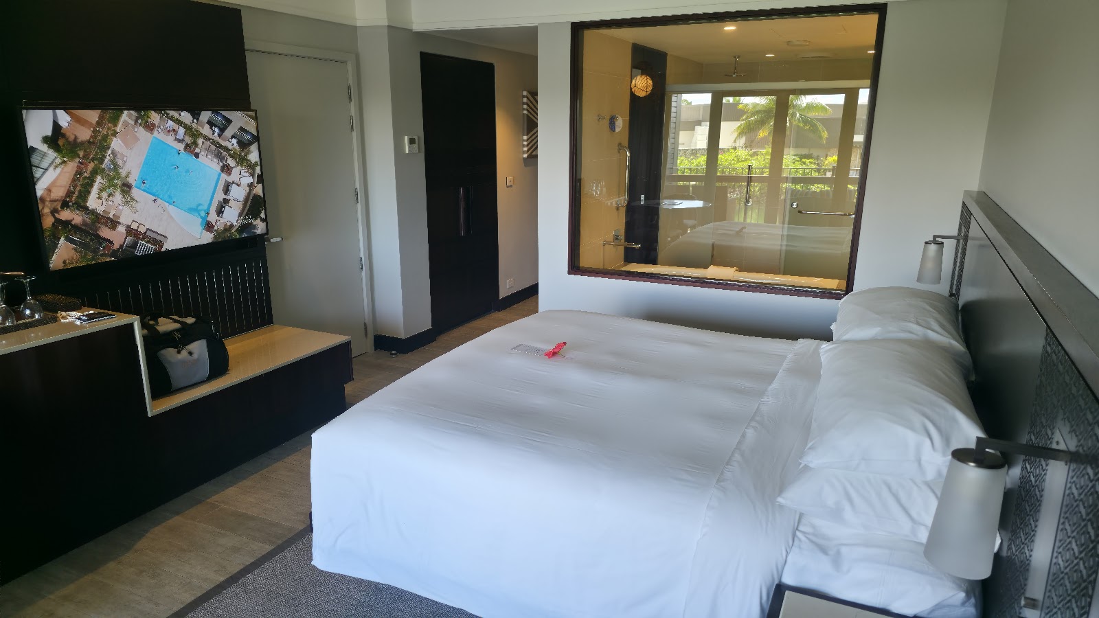 Modern resort guest room with two white beds, flat-screen TV displaying aerial pool view, and glass doors opening to a balcony with tropical greenery.