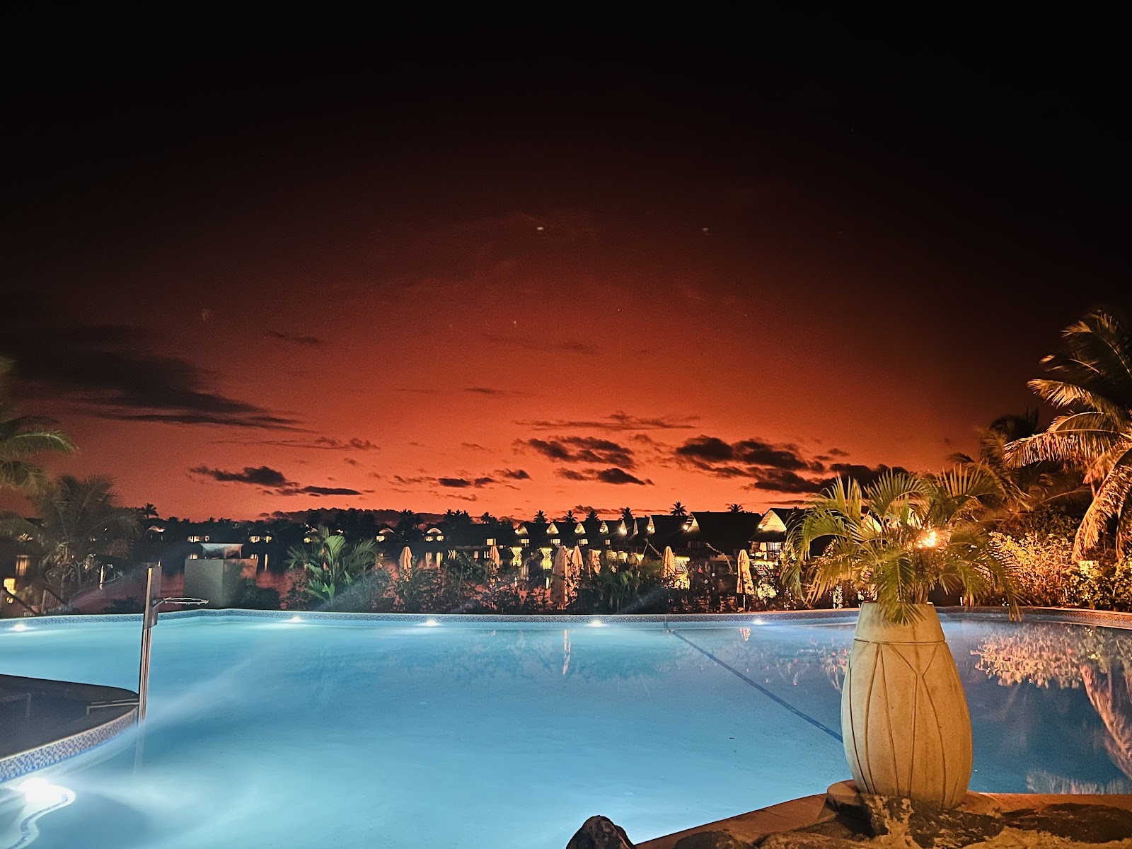 Illuminated pool at sunset with palm trees, resort bungalows, and dramatic orange sky reflected in the water.