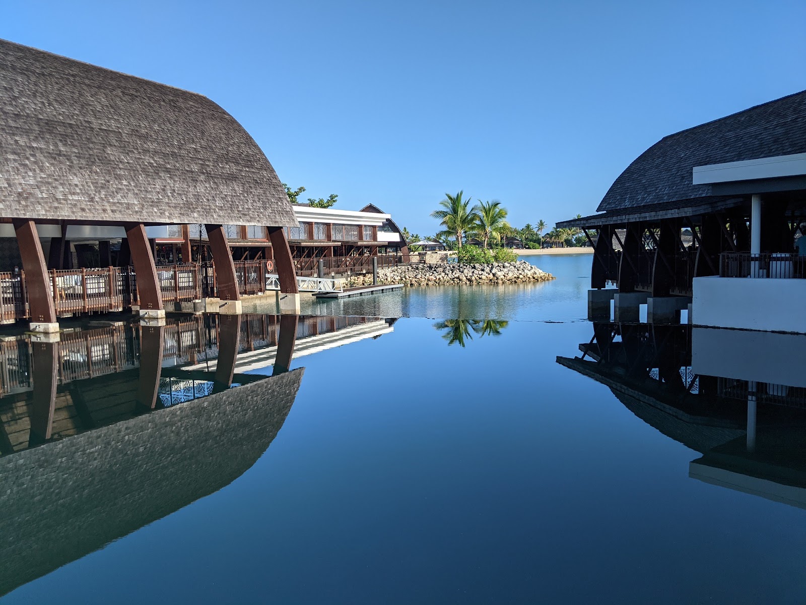 Thatched-roof overwater bungalows with brown supports line a calm lagoon at Fiji Marriott Resort Momi Bay, with palm trees and rocky shore beyond.