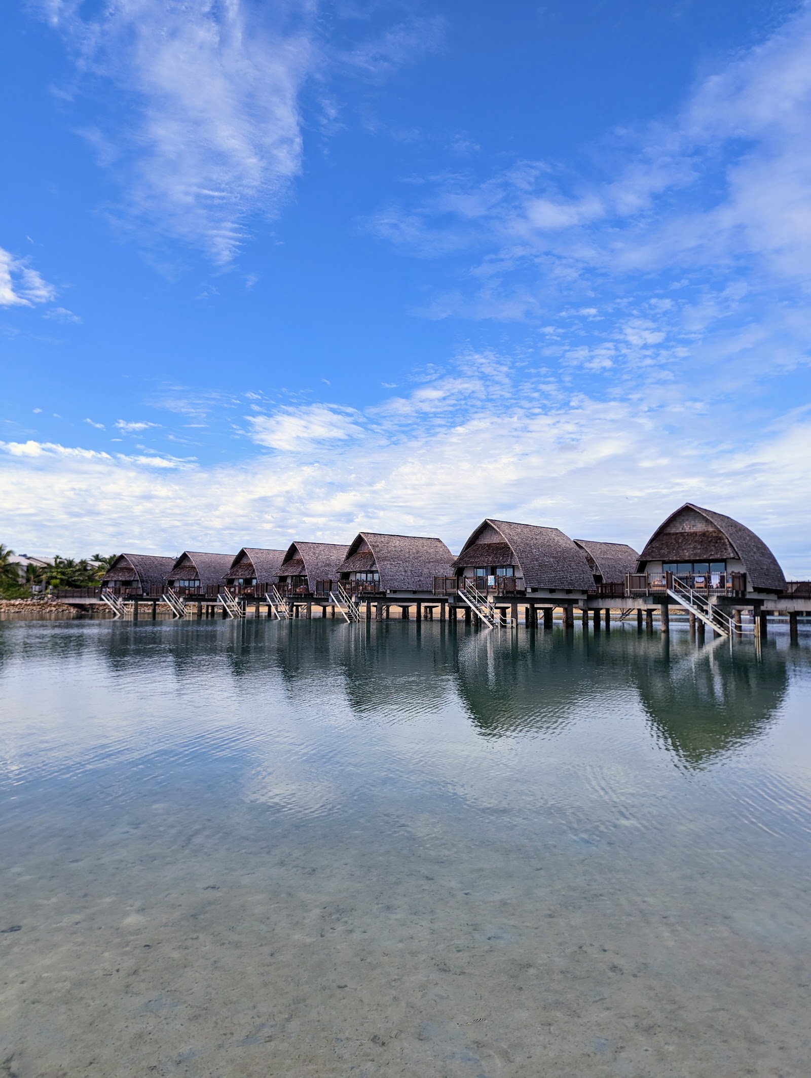 Row of thatched-roof overwater bungalows on stilts at Fiji Marriott Resort Momi Bay, with shallow turquoise water and sandy beach in foreground.