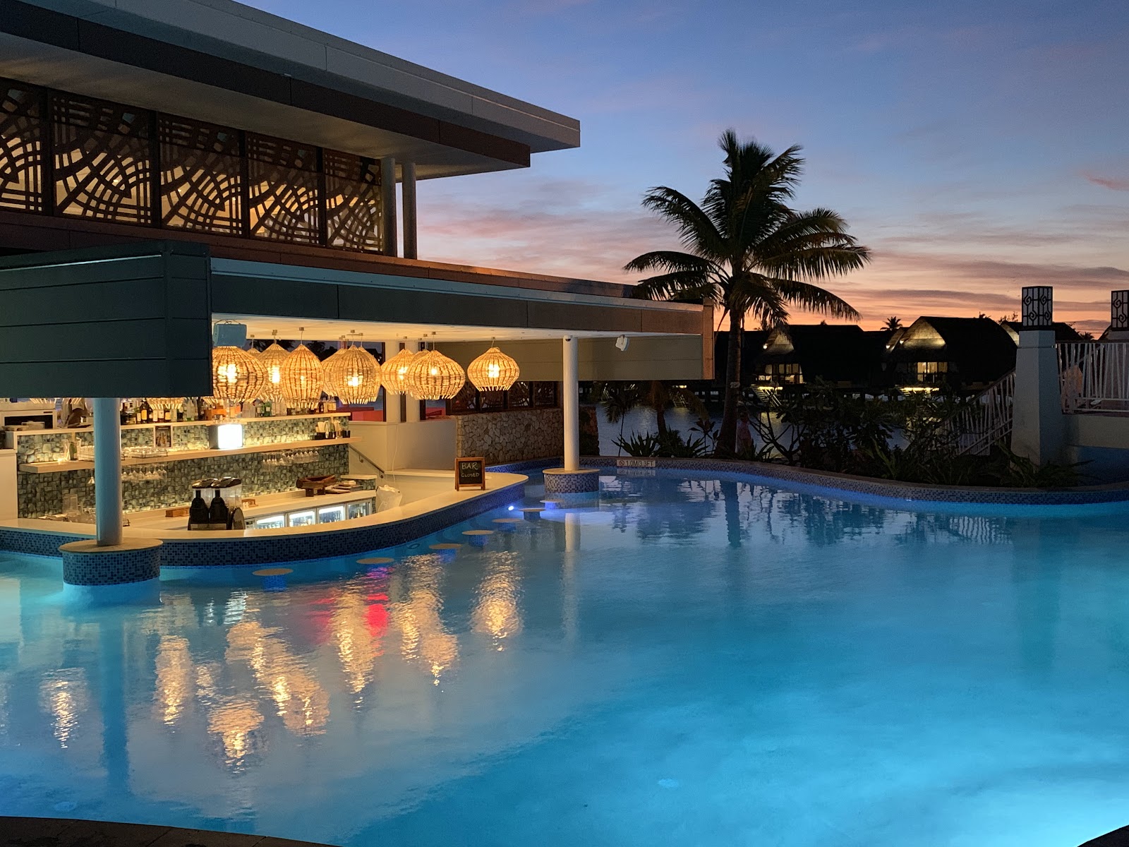 Poolside bar with woven pendant lights at dusk, turquoise water reflecting the sunset sky, palm trees and thatched-roof bungalows visible beyond.