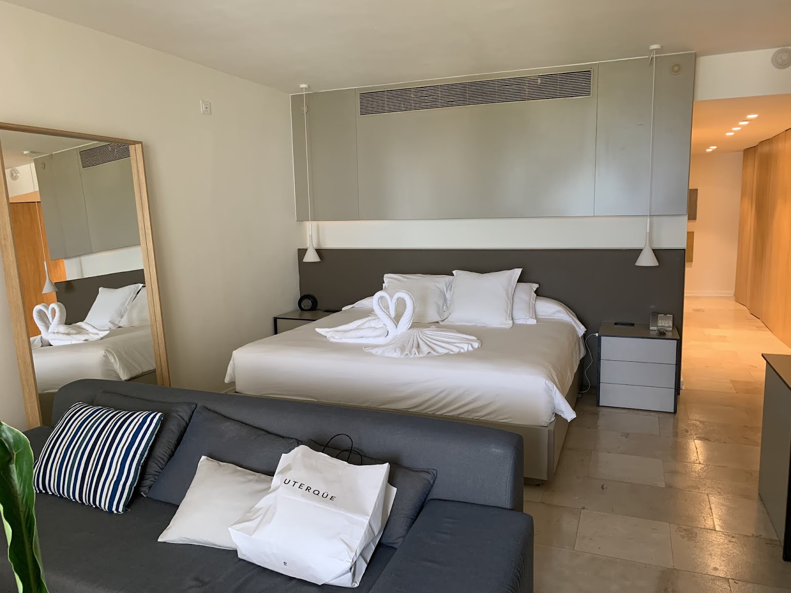 Modern resort bedroom with king bed, white linens, decorative towel swan, dark gray headboard, sitting area with dark sofa, and mirror-lined wall at Finest Playa Mujeres.