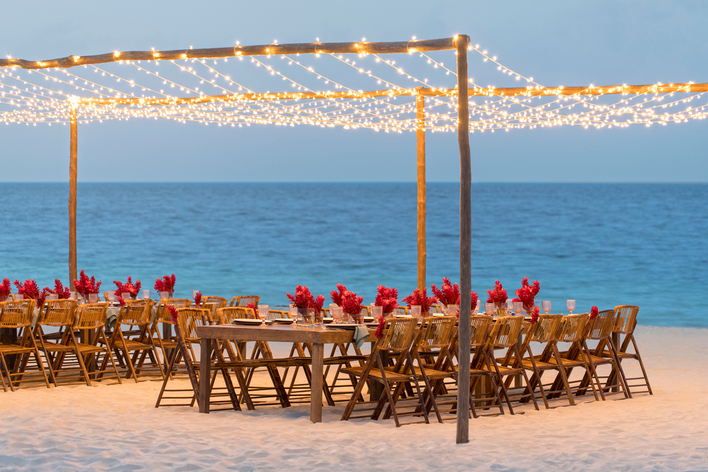 Beachfront dining setup at Finest Playa Mujeres with wooden chairs, red flower centerpieces, string lights overhead, and ocean view.