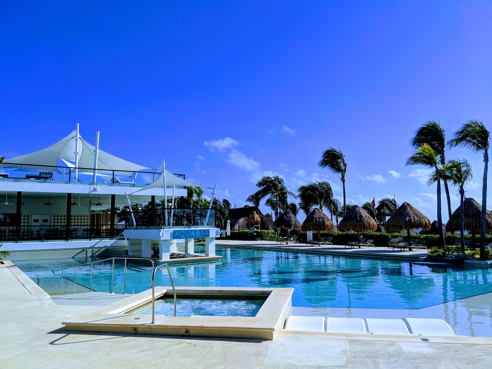 Resort pool with Aqua Bar, thatched palapas, palm trees, and clear blue sky at Finest Playa Mujeres.