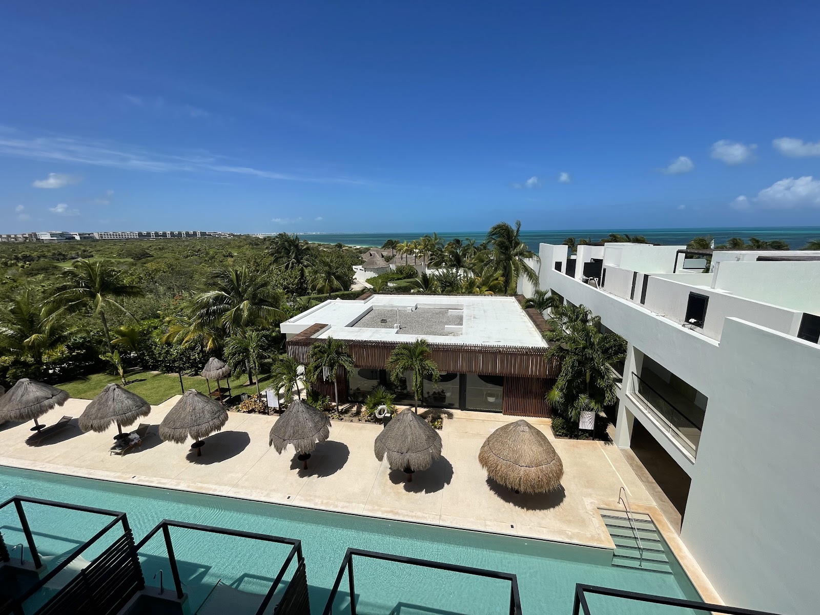 Elevated view of Finest Playa Mujeres pool area with thatched umbrellas on sand, modern white buildings, palm trees, and turquoise ocean beyond.