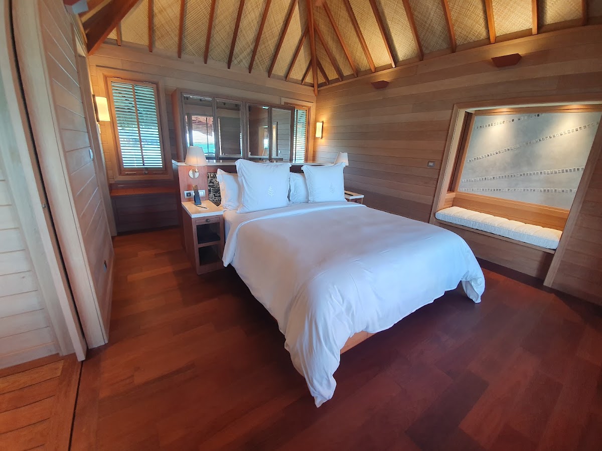 Overwater bungalow bedroom with wooden interior, pitched ceiling, queen bed with white linens, and ocean view through glass doors.