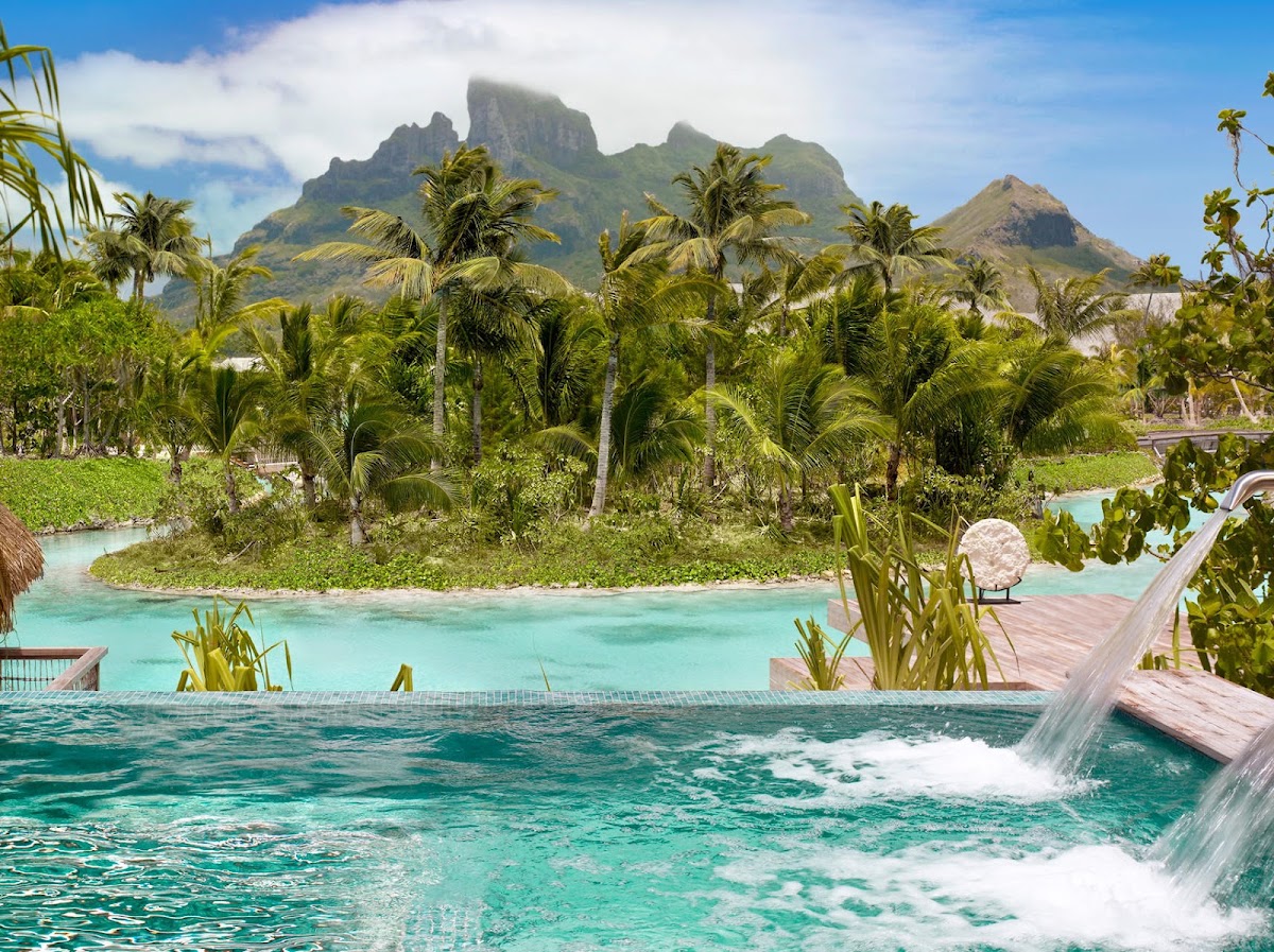 Turquoise lagoon with wooden deck, palm trees, and dramatic mountain peaks rising behind lush vegetation.