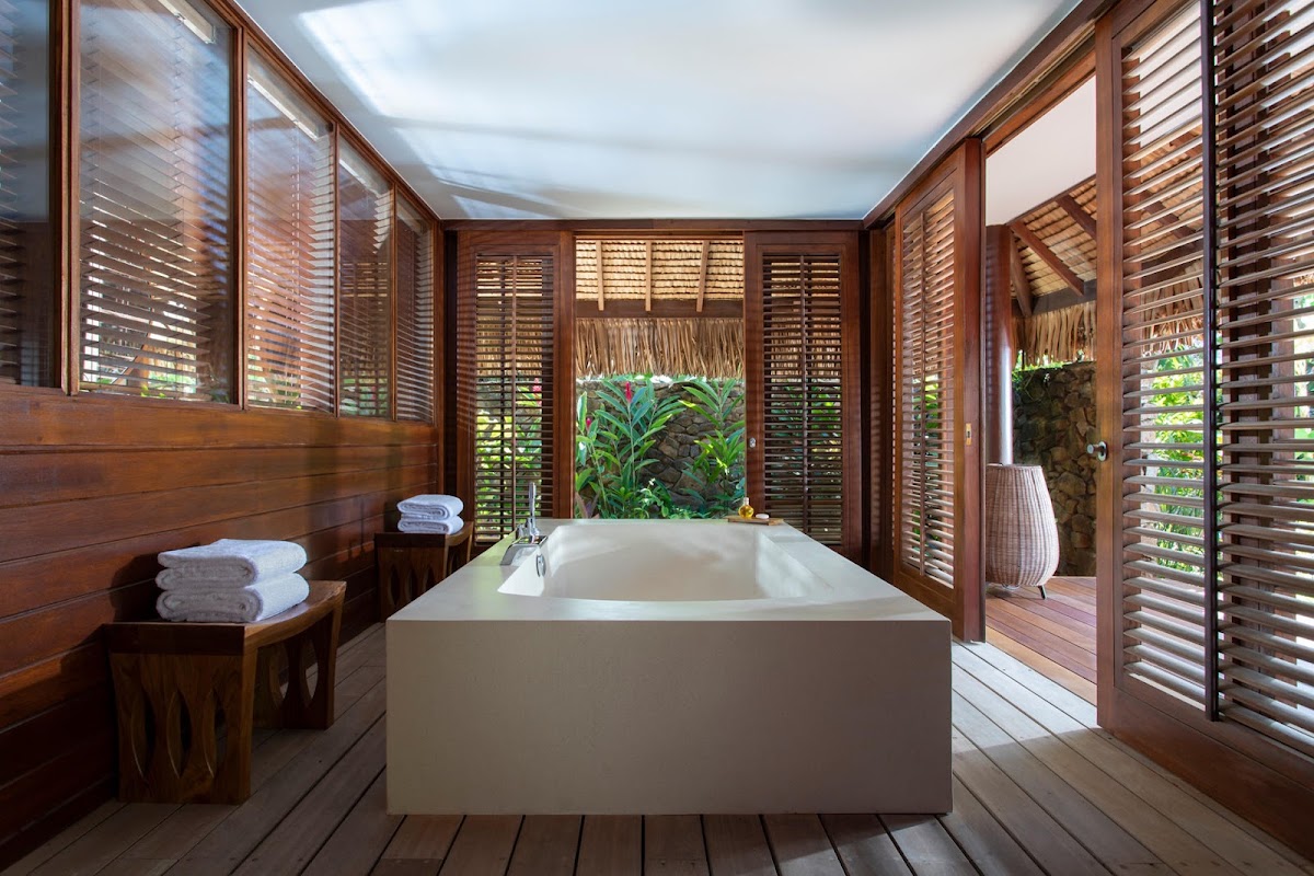 Open-air bathroom at Four Seasons Bora Bora with white soaking tub, wooden shutters, tropical plants, and towels on a side table.