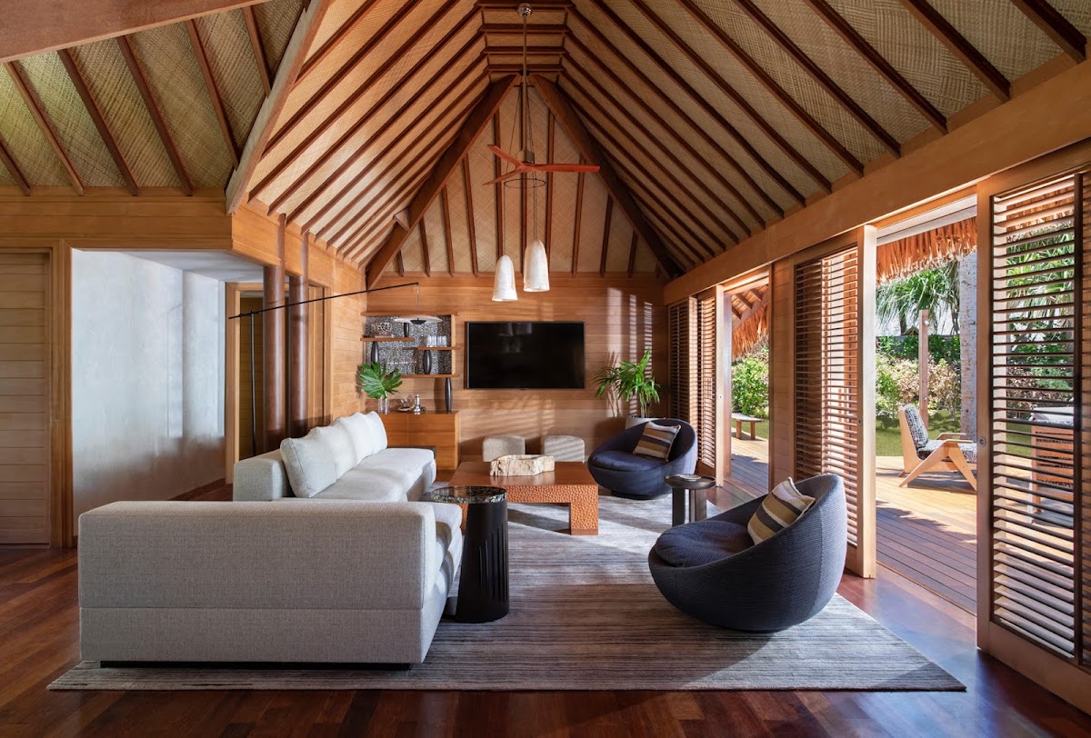 Four Seasons Bora Bora overwater bungalow interior with wooden vaulted ceiling, white sectional sofa, black curved chairs, and louvered doors opening to a deck and lagoon views.
