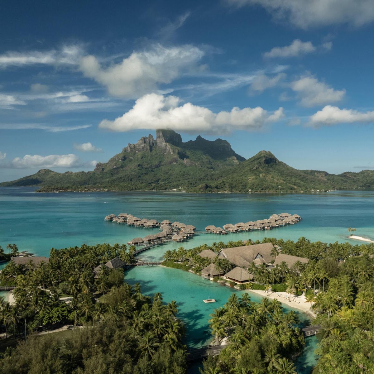 Four Seasons Bora Bora lagoon view with overwater bungalows, turquoise water, palm trees, and volcanic peaks in the distance.