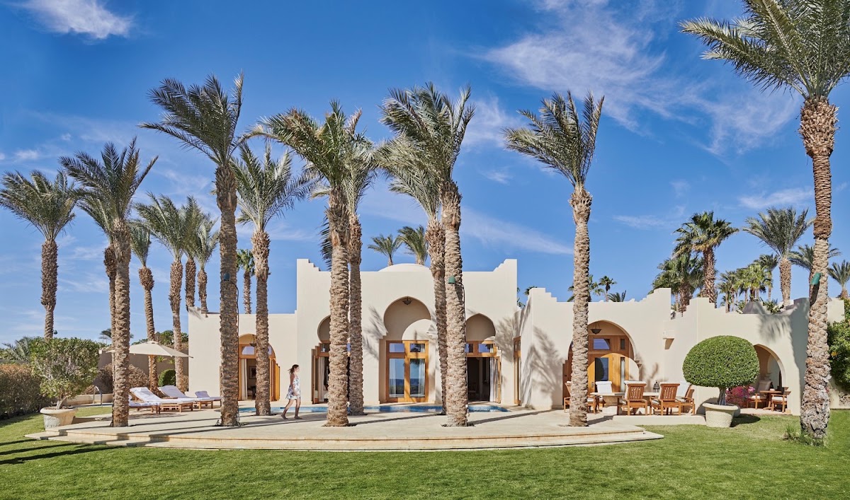 White curved building with arched doorways and wooden doors surrounded by tall palm trees, lounge chairs, and manicured grass under clear blue sky.