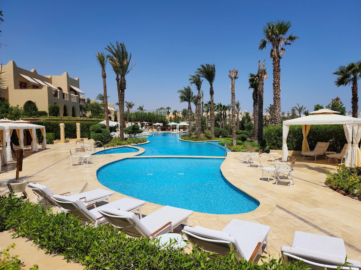 Resort pool area with curved blue water, lounge chairs with white cushions, cabanas, palm trees, and beige colonial-style buildings under clear sky.