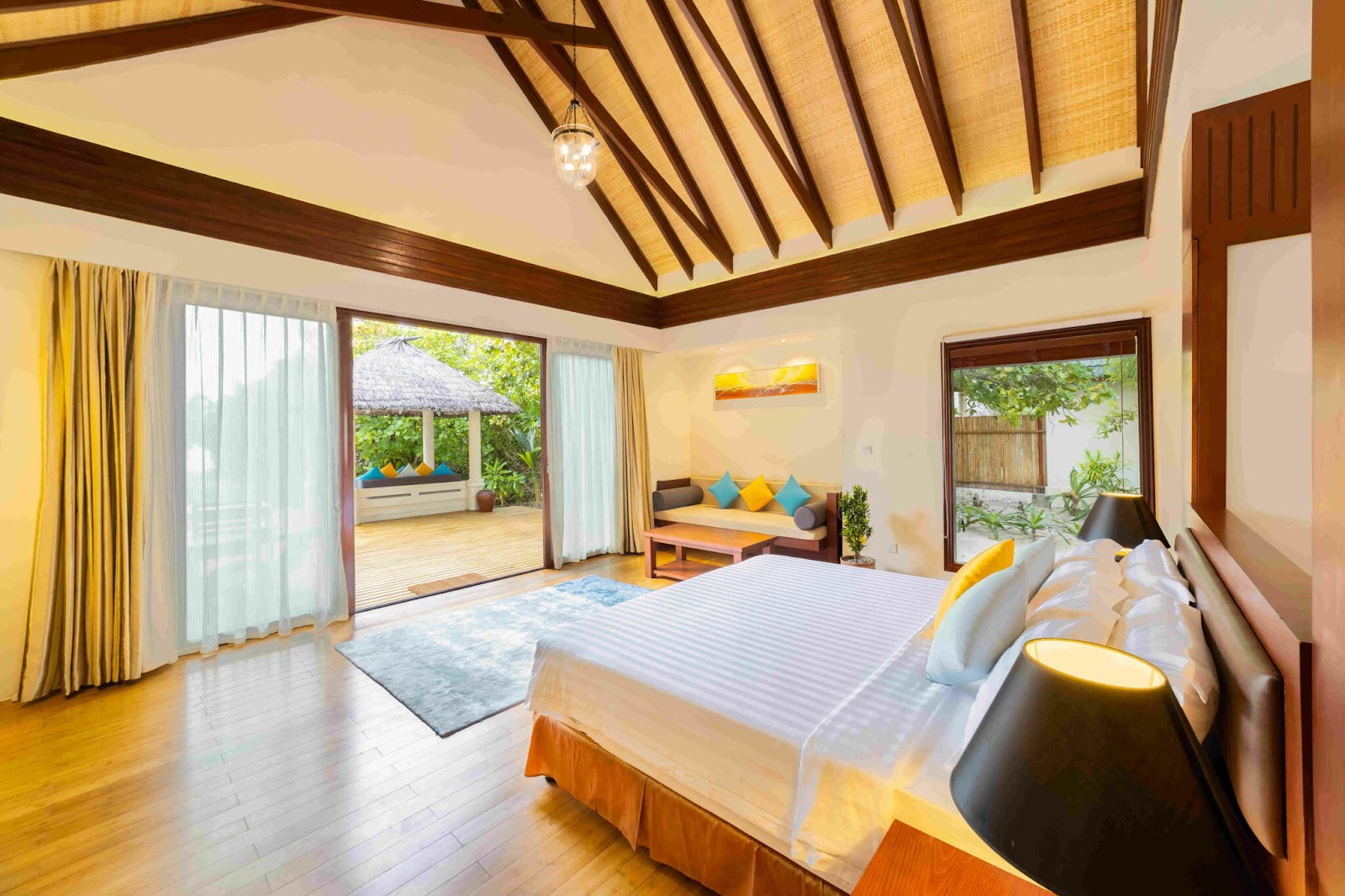 Spacious villa bedroom with wooden beams, striped bed, and open doors leading to a furnished patio with thatched roof and ocean views.