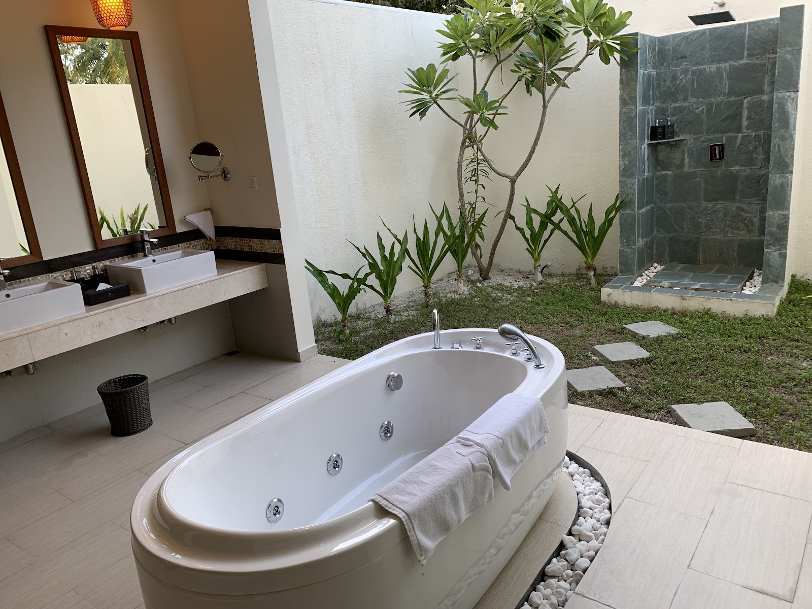 Outdoor bathroom with sunken oval soaking tub, vanity with mirror, and open-air shower surrounded by tropical plants and white pebbles.