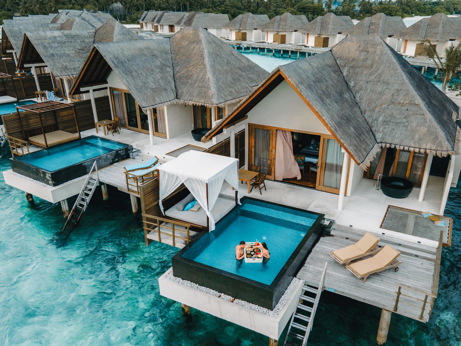 View from an open bungalow through wooden shutters to a tropical garden path with thatched-roof villas and lush vegetation.