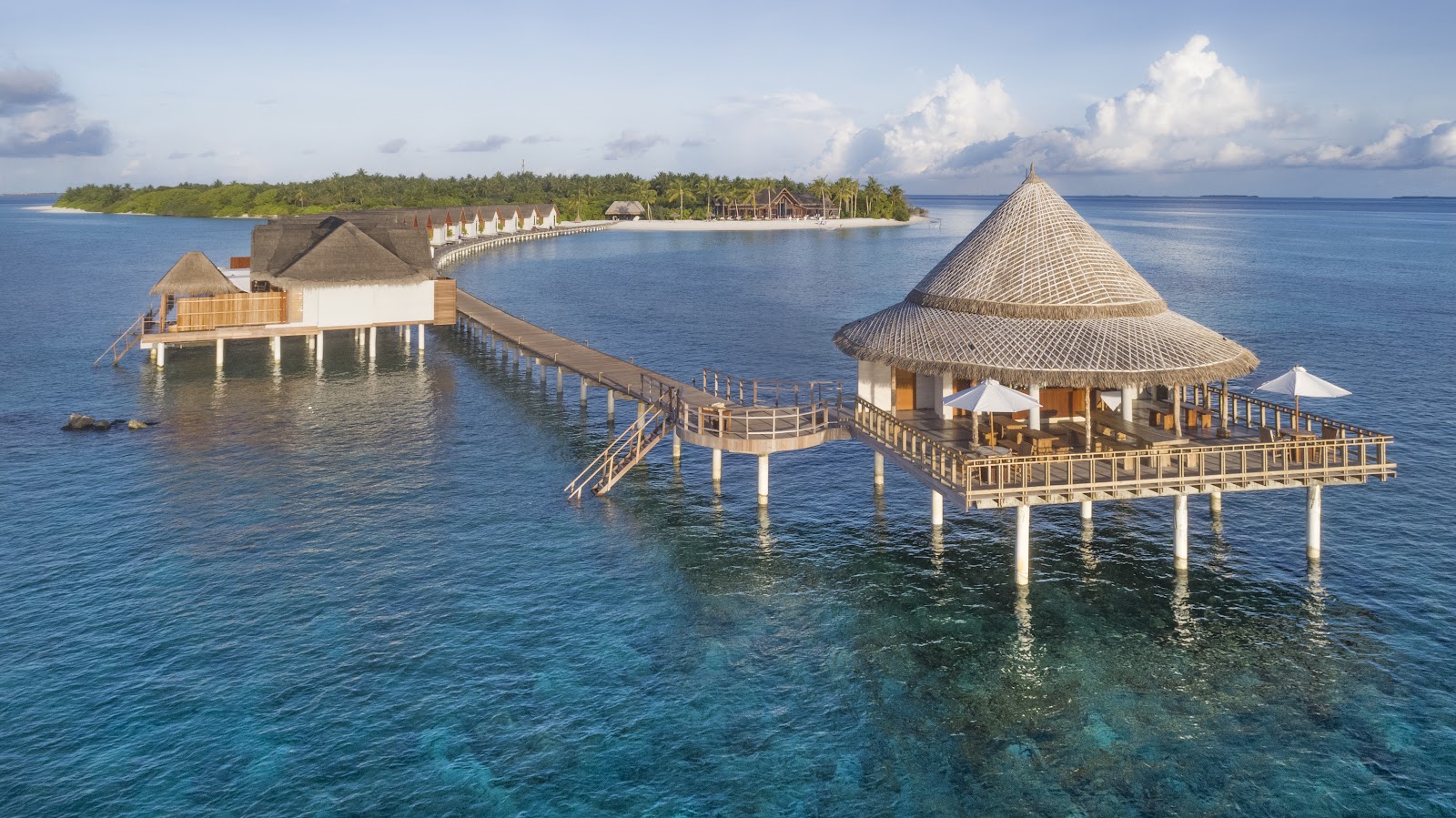 Aerial view of Furaveri Maldives nestled in dense palm forest with turquoise lagoon, blue pool, white sand beach, and thatched bungalows along a stepped hillside.