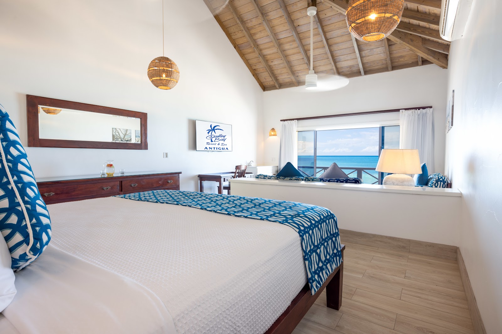 Beachfront bedroom at Galley Bay Resort & Spa with ocean views, blue patterned bedding, woven pendant lights, and wooden beam ceiling.