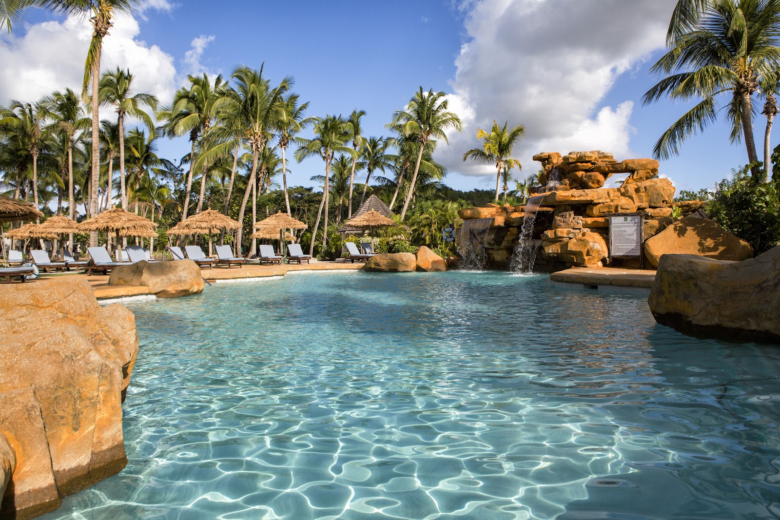 Turquoise lagoon with sandy bottom surrounded by large boulders, palm trees, and thatched umbrellas with lounge chairs at Galley Bay Resort & Spa.
