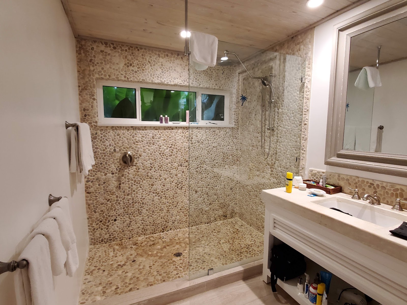 Bathroom with pebble-tiled walk-in shower, glass enclosure, double vanity with mirror, and wood ceiling.