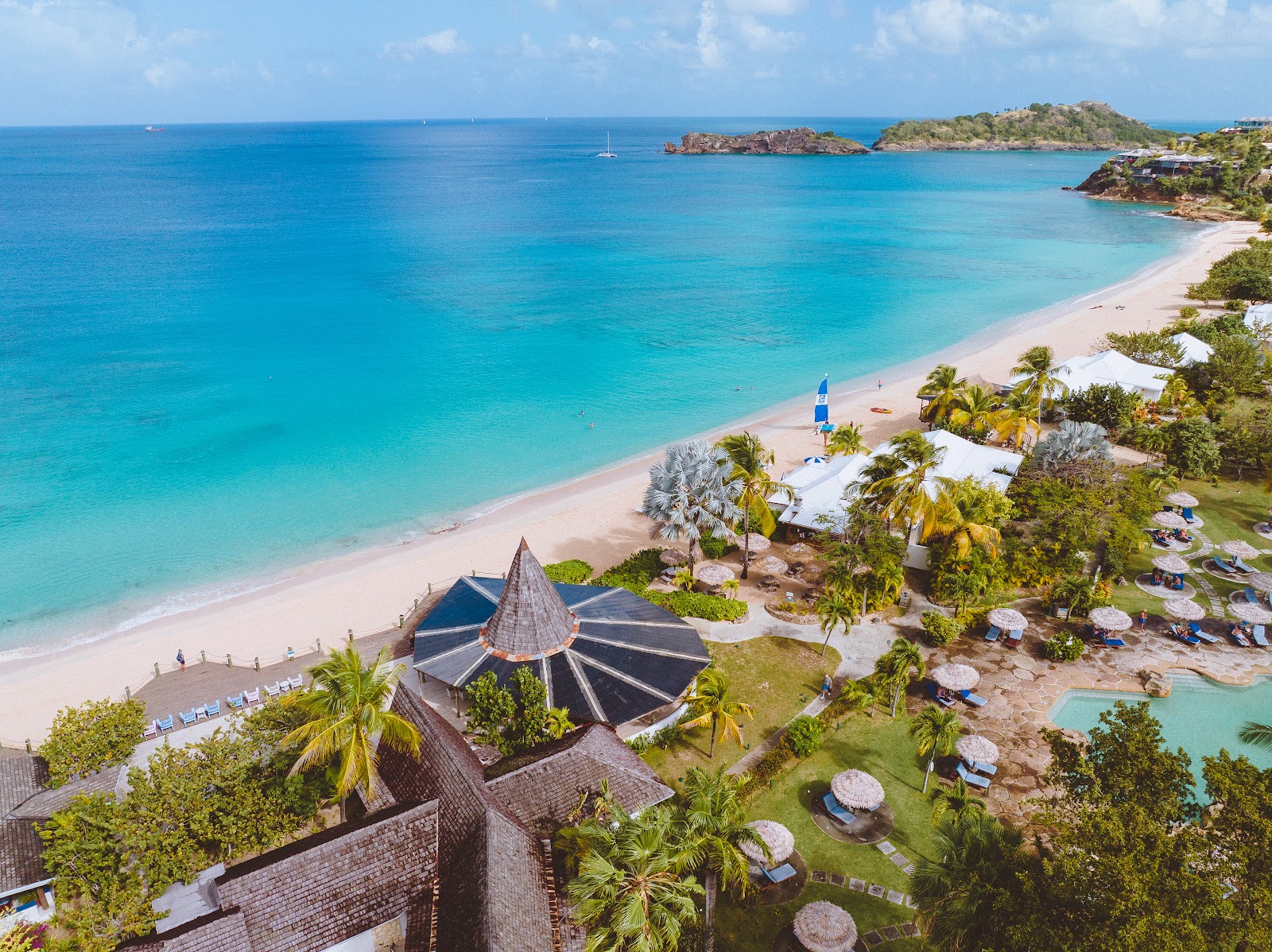 Aerial view of Galley Bay Resort & Spa's beach and grounds with turquoise water, pink sand, and thatched-roof structures surrounded by palms.