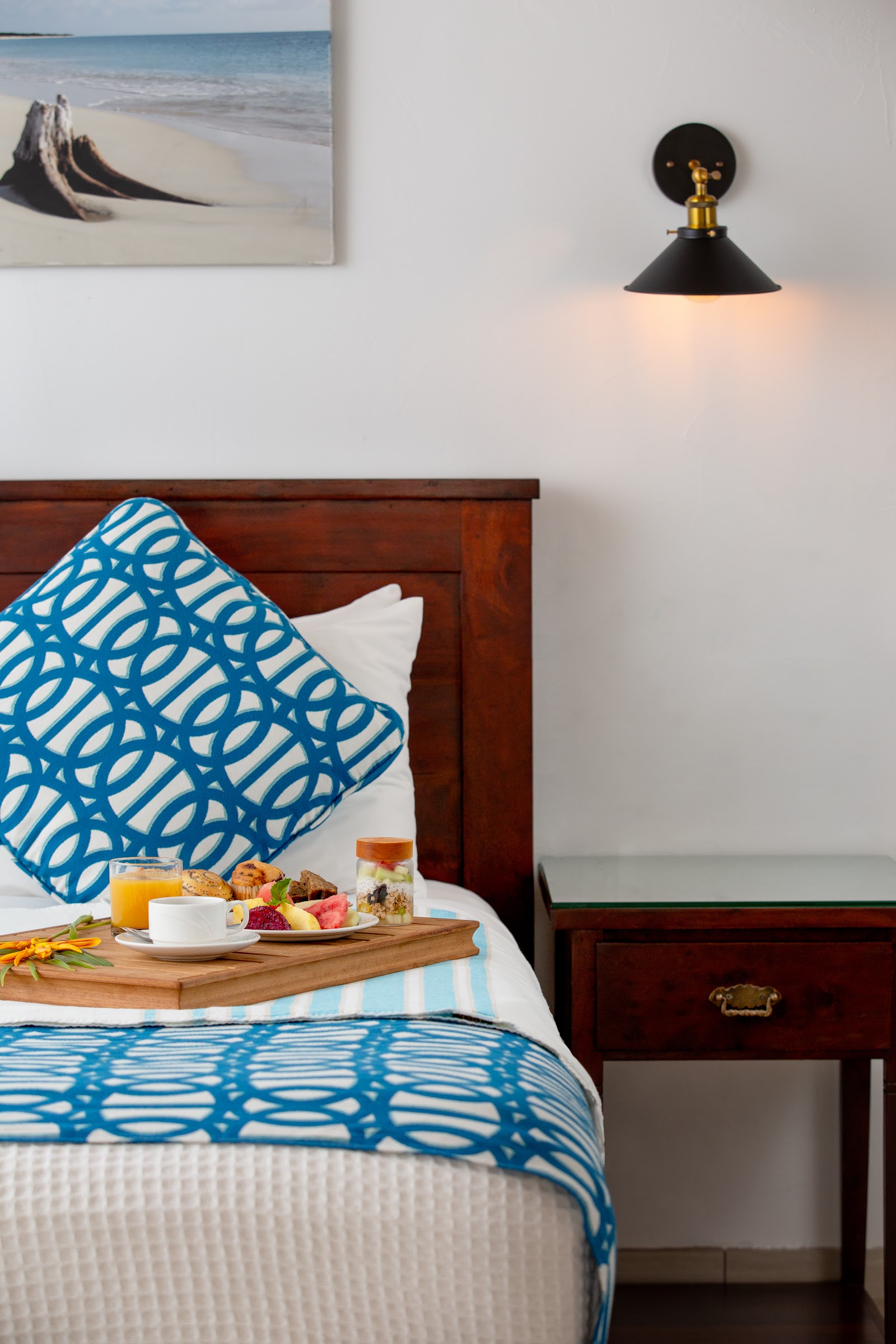 Hotel room with wood bed frame, blue patterned bedding, breakfast tray with fruit and juice, and beach photograph on wall.