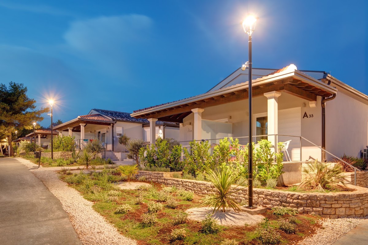 Row of white cottages with porches lit by streetlamps at dusk, surrounded by Mediterranean plants and gravel landscaping.