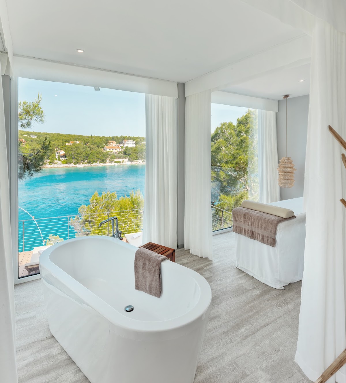 Bathroom with white oval freestanding tub overlooking turquoise Adriatic Sea, green coastline, and hillside villas through floor-to-ceiling windows.