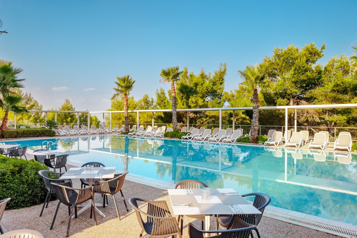 Large turquoise pool at Gava Waterman Milna Resort surrounded by white loungers, dining tables, and palm trees under clear blue sky.