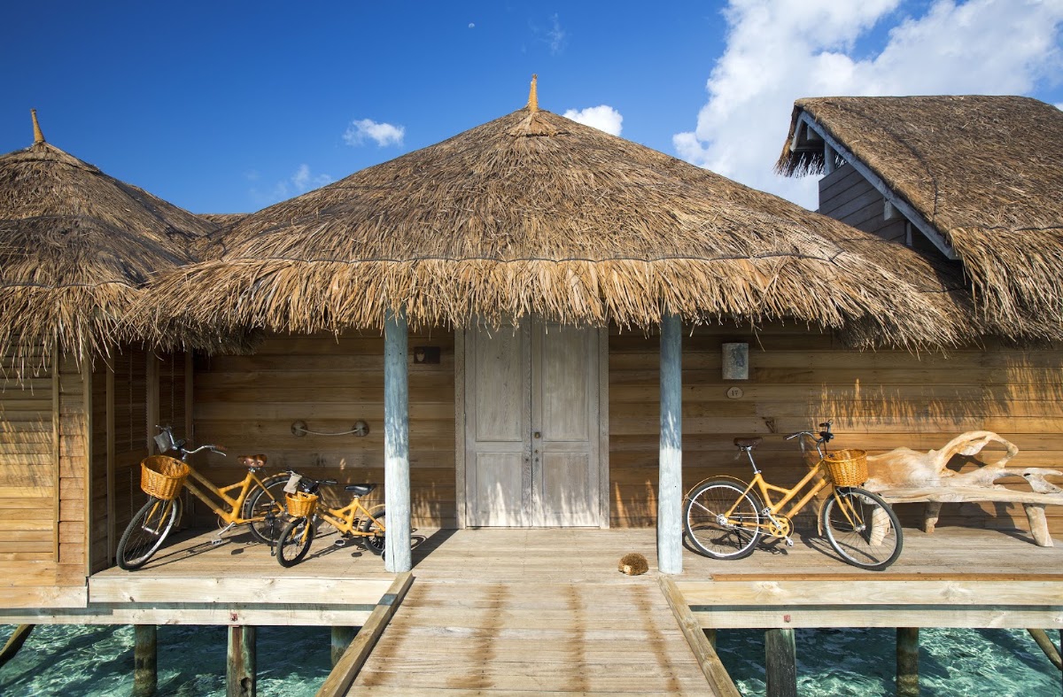 Overwater bungalows with thatched roofs and yellow bicycles on wooden decks at Gili Lankanfushi, with clear blue sky above.