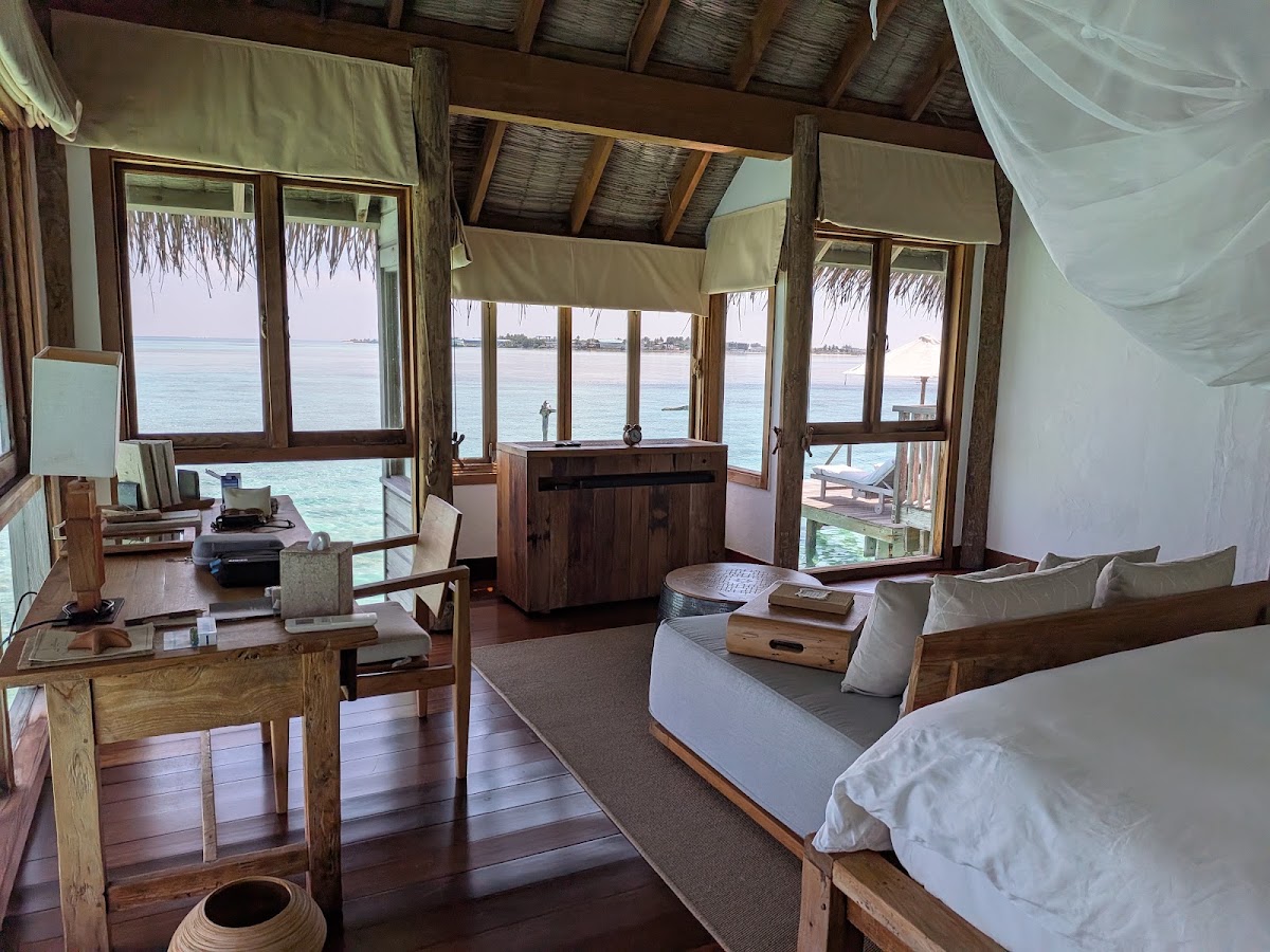 Overwater bungalow interior at Gili Lankanfushi with wooden furnishings, bed, desk, and expansive windows overlooking turquoise lagoon and distant islands.