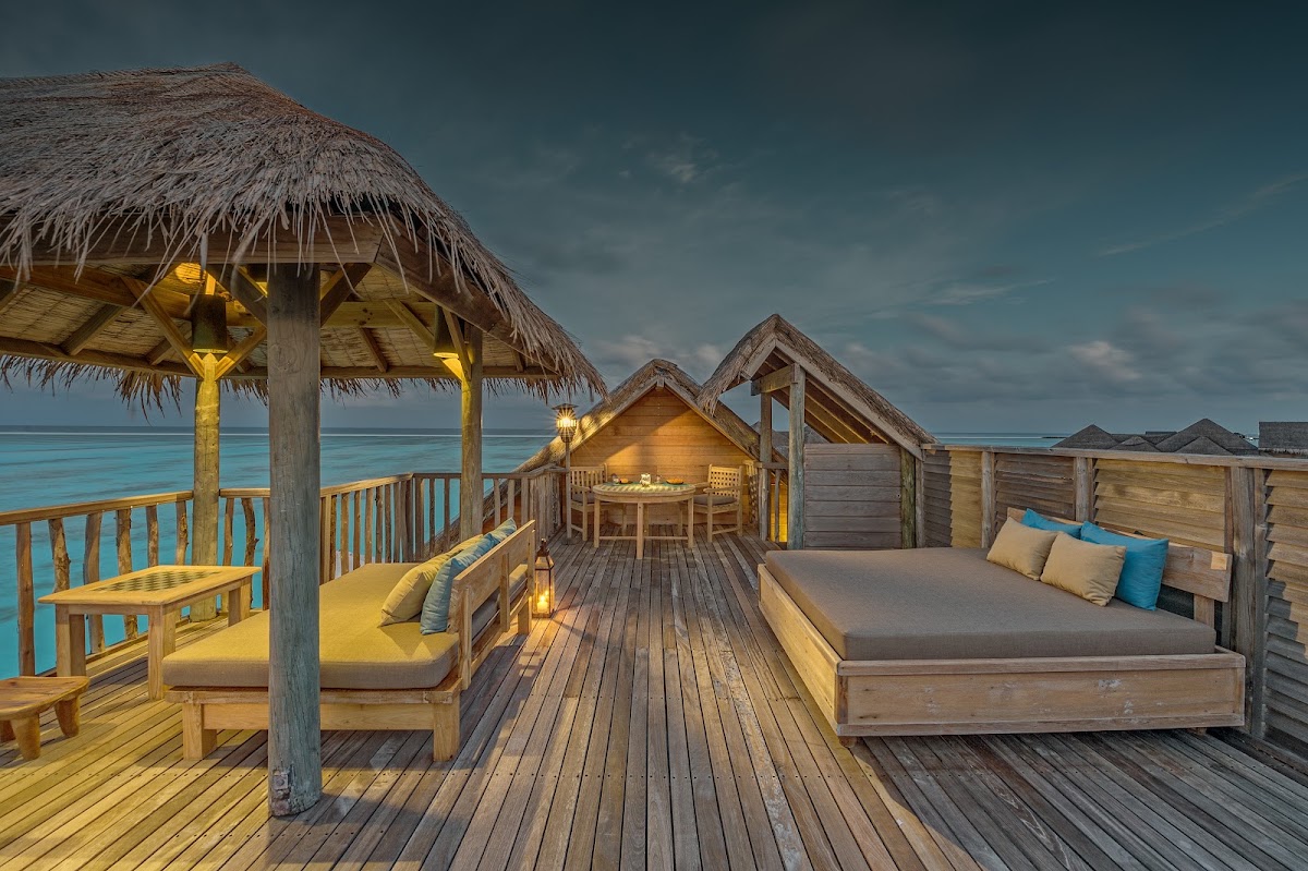 Wooden overwater bungalows with thatched roofs and lit interiors line a deck at Gili Lankanfushi, with turquoise ocean and sky beyond.