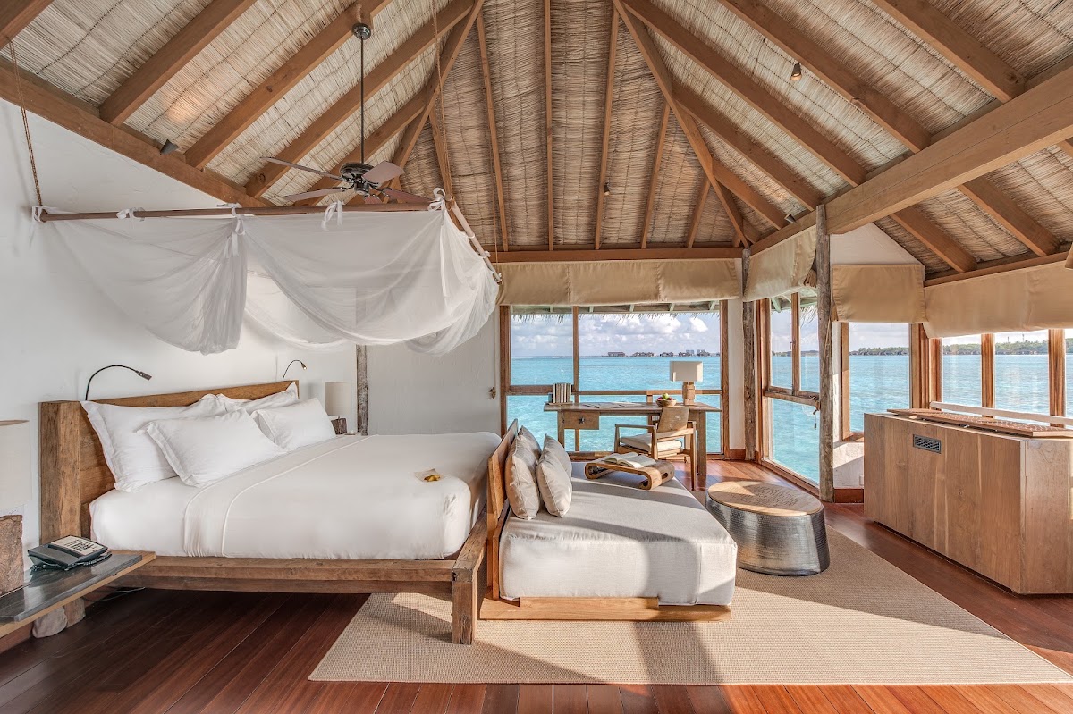 Overwater bungalow interior at Gili Lankanfushi with wooden bed, white linens, woven ceiling, and turquoise ocean views through glass doors.