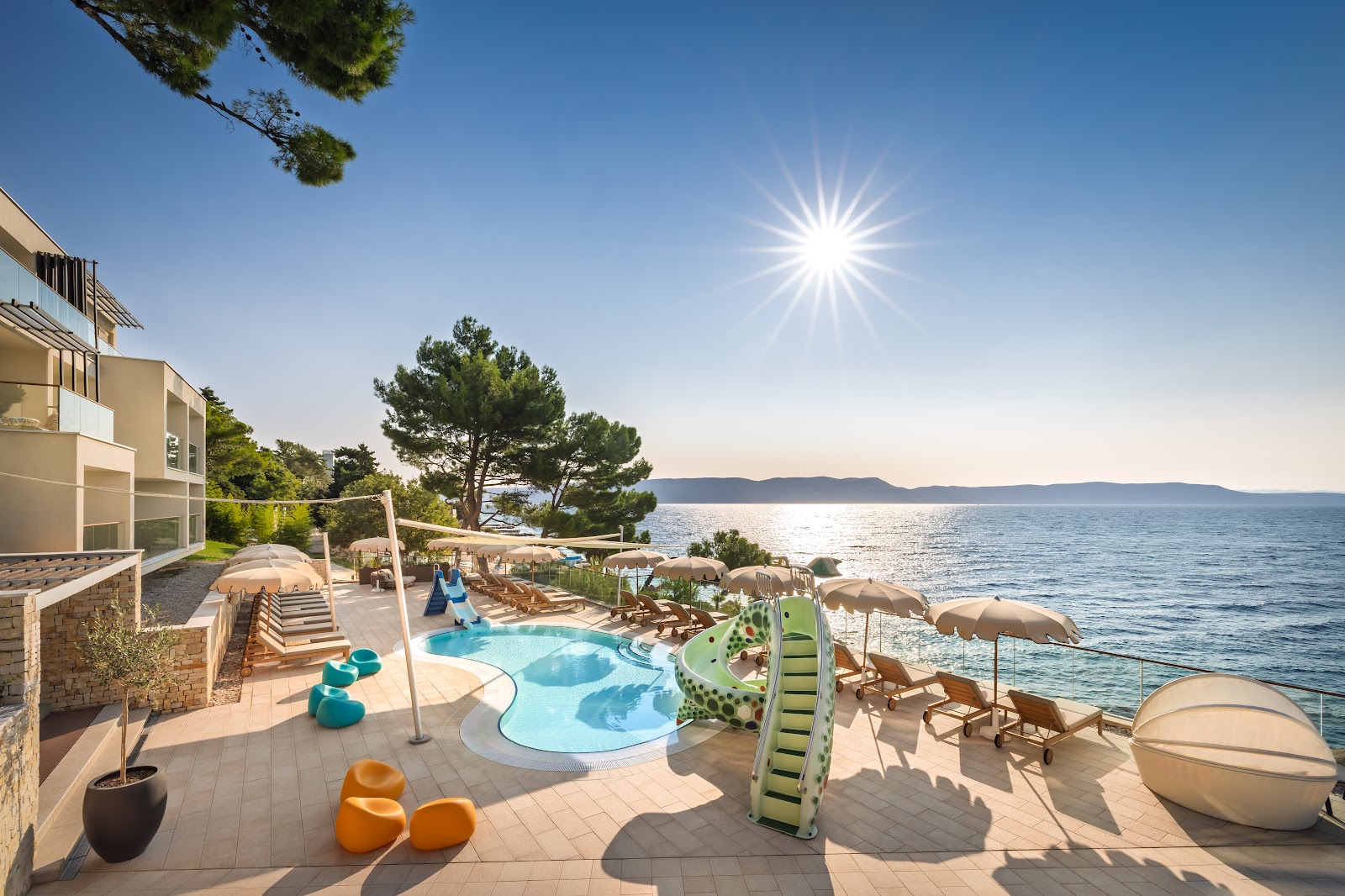 Waterfront resort pool area with curved swimming pool, lounge chairs under umbrellas, beige buildings, and Adriatic Sea with distant mountains under bright sunshine.