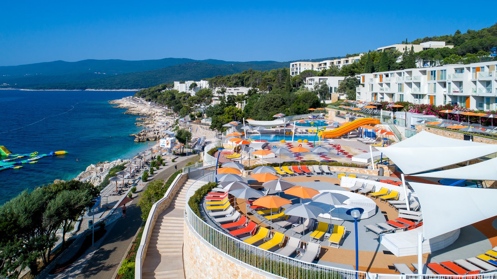 Girandella Resort's pool area with colorful umbrellas and loungers overlooks a rocky Adriatic coastline and forested hillside under clear blue sky.